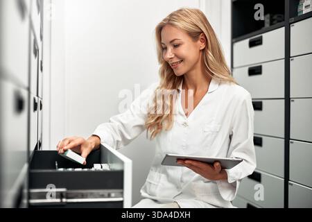 Smiling female pharmacist in white coat using digital tablet while checking medication in storage drawers. Modern pharmacy concept. Stock Photo