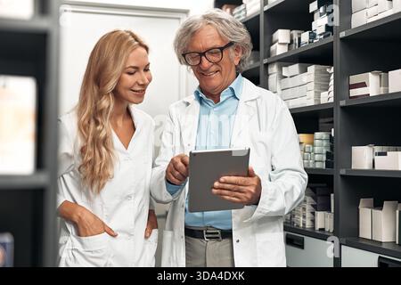 Pharmacists standing between shelves, using tablet to review medicine stock. Collaboration in pharmaceutical storage. Concept: healthcare, inventory, Stock Photo