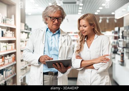Senior male pharmacist showing digital information to young female colleague in modern pharmacy. Professional teamwork in healthcare concept. Stock Photo