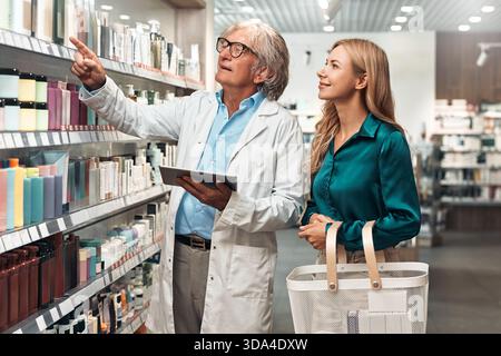 Smiling female customer with shopping basket listens to pharmacist explaining product options in a modern pharmacy aisle. Concept: healthcare, consult Stock Photo