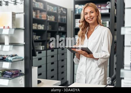 A young female pharmacist in a white coat checking stock using a digital tablet in a modern pharmacy. Digital tools for healthcare management. Stock Photo