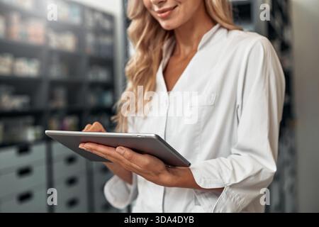 A young female pharmacist in a white coat checking stock using a digital tablet in a modern pharmacy. Digital tools for healthcare management. Stock Photo