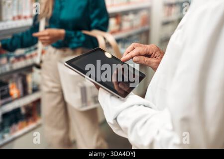 Senior pharmacist using a digital tablet in a pharmacy, assisting a customer with product selection. Modern pharmacy with cosmetic and skincare shelve Stock Photo