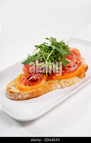 Bruschetta featuring prosciutto, sliced tomatoes, and fresh arugula on a white plate Stock Photo