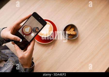 Person holding smartphone, capturing content of latte art coffee and biscotti on table Stock Photo