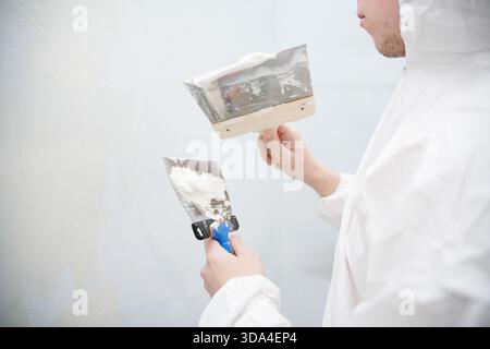 Worker wearing a protective suit is plastering a wall using two putty knives, restoring a room Stock Photo