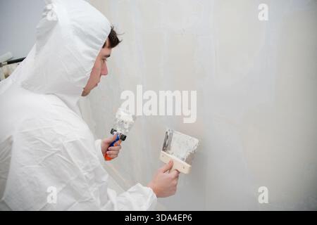 Man wearing protective suit applying plaster to a wall with spatulas Stock Photo