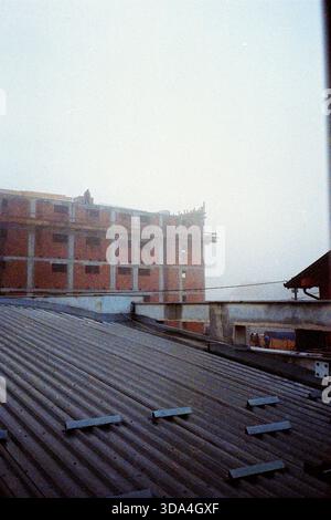 Grainy analog shot of a red block building under construction in winter fog, with one worker on the roof and single-story houses in the foreground. Stock Photo