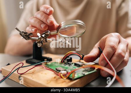 Close-up hands of technician focusing on intricate details of circuit board on wooden base with magnifying glass, using helping hands tools and wires Stock Photo