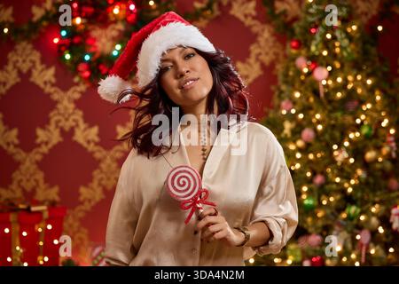 Woman wearing Santa hat holds candy cane lollipop in front of decorated Christmas tree Stock Photo