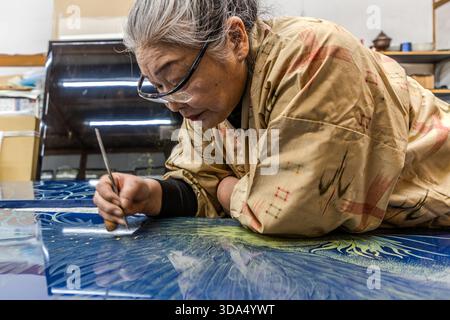 An artist carving a blue lacquer panel – plant motifs and curving lines in white and green take shape; in the background, shelves displaying finished lacquerwork. Shiojiri, Nagano Prefecture, Japan Stock Photo
