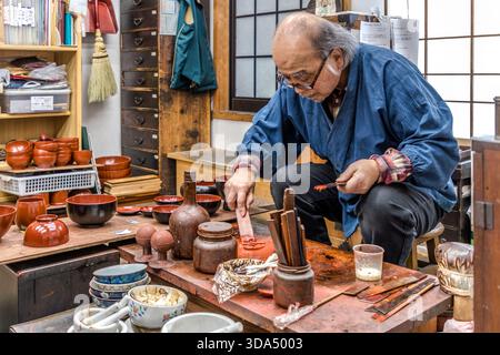 An urushi lacquer craftsman in his traditional workshop, mixing red urushi lacquer on the worktop; in the background, shelves lined with numerous finished red wooden bowls of various sizes. Shiojiri, Nagano Prefecture, Japan Stock Photo