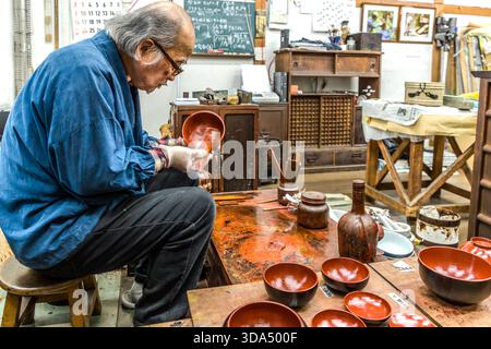 A lacquer craftsman in his urushi workshop, examining a bowl; on the workbench and in the background are dozens of wooden bowls at various stages of production – from the primer coat to the finished red lacquer surface. Shiojiri, Nagano Prefecture, Japan Stock Photo