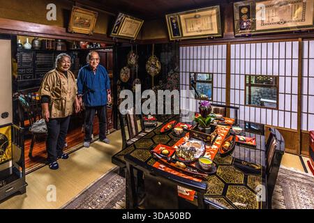 A Japanese couple stand in the festively laid tatami room of a traditional guesthouse (minshuku) in the Kiso region, Nagano Prefecture. The lacquered wooden table is set with several bowls and trays for a kaiseki-style dinner; there are shoji sliding doors and framed certificates on the wall. Such family-run establishments are still common today along the old Nakasendo Post Road in the Kiso Valley. Shiojiri, Nagano Prefecture, Japan Stock Photo