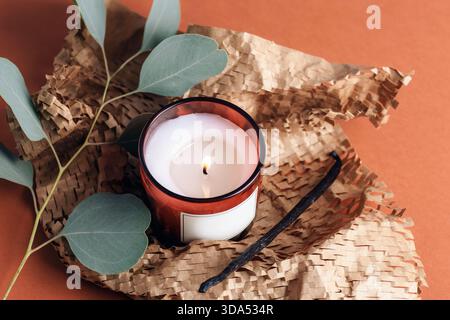 Lit candle in red glass with eucalyptus leaves and textured paper on warm orange background. Top view. Stock Photo