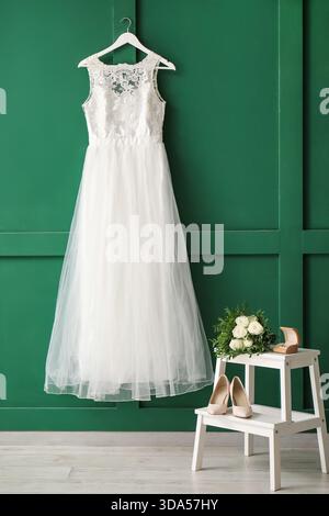 Beautiful wedding dress on green wall with ring box, wedding shoes and bridal bouquet on white wooden stepladder Stock Photo