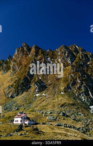 Isolated alpine house, mountain cabin beneath dramatic rocky peaks in Fagaras Mountains, Romania. Stock Photo