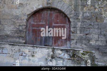blurred rustic arched wooden door set in old stone castle wall, viewed from behind a textured, weathered foreground concrete ledge with exposed rebar Stock Photo