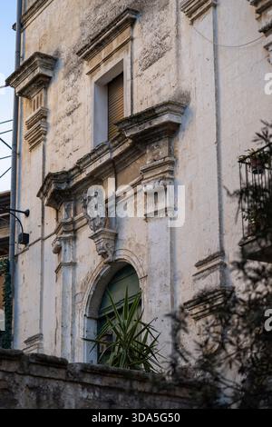 baroque architecture detail of an ancient, weathered stone building facade in southern italy, featuring ornate carvings, an arched window, and a plant Stock Photo