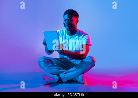 Young Man Enjoys Digital Content While Sitting on the Floor in Colorful Lighting Stock Photo