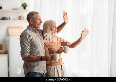 Elderly Couple Enjoying Morning Coffee and Sharing a Moment Together While Waving at Someone Outside Their Bright Kitchen Stock Photo