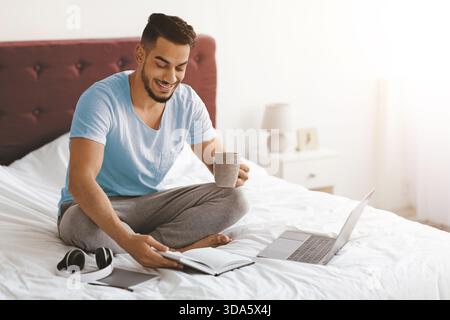 Man Enjoying Morning Routine While Sitting on Bed With Laptop and Notebook Stock Photo