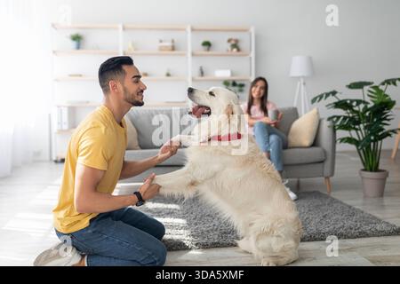 Young Man Plays With Golden Retriever Dog in Cozy Living Room While Woman Relaxes on Couch Stock Photo