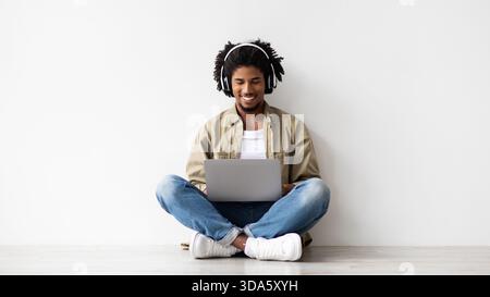 Young African American Guy Working on Laptop With Wireless Headphones While Sitting on Floor Stock Photo