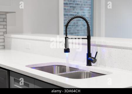 Close up of a modern kitchen sink with a black pull down faucet featuring blue LED lights, grey cabinets, and a white quartz countertop. Stock Photo