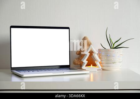 Laptop with blank screen on desk with Christmas decorations, small pine tree candle holder, and potted plant with string light for holiday season. Stock Photo