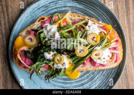 Appetizing lunch. Portion dish. Sandwich with vibrant toppings serving on plate. Food with green grilled leaves beetroot olives yolk sauce. Stock Photo