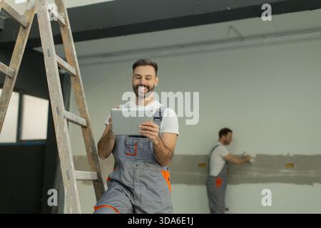 Two workers are engaged in a renovation project. One is using a tablet while standing on a ladder, while the other is applying plaster on the wall. Stock Photo