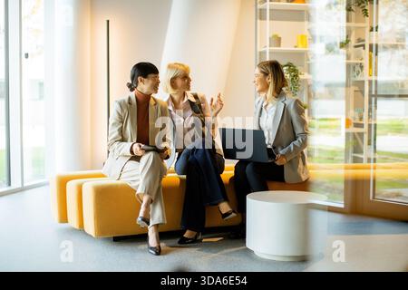 Three businesswomen engage in a lively discussion in a contemporary office lounge. They are seated on a cozy bench, sharing ideas and looking at a lap Stock Photo