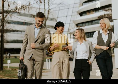 Four professionals stroll together in a vibrant business area, engaging in conversation and enjoying the pleasant weather. Their attire reflects a mod Stock Photo