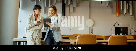 Two professionals are discussing work-related topics in a contemporary office. They are engaged while reviewing information on a tablet at a shared wo Stock Photo