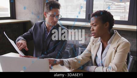 Discussing two colleagues pointing at laptop and holding document in modern office, wearing blazers Stock Photo