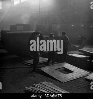 Slavyansk, Ukrainian SSR, USSR - 1970s: Three Soviet workers having a conversation in the smoky atmosphere of the Stroymash factory workshop. The men in workwear are standing next to a huge steel cylinder section, taking a short break from heavy labor. Bright sunbeams cut through the thick industrial haze, highlighting the metal plates scattered on the floor. This black and white archival photo captures the authentic daily life and tough working conditions of the peaceful Donbass region during the Soviet era. Men in heavy industry. Stock Photo