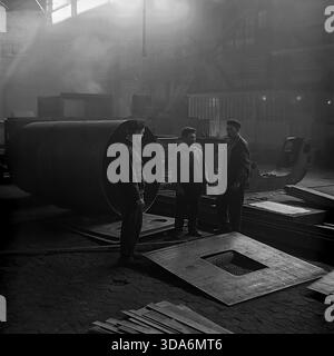 Slavyansk, Ukrainian SSR, USSR - 1970s: Three Soviet workers having a conversation in the smoky atmosphere of the Stroymash factory workshop. The men in workwear are standing next to a huge steel cylinder section, taking a short break from heavy labor. Bright sunbeams cut through the thick industrial haze, highlighting the metal plates scattered on the floor. This black and white archival photo captures the authentic daily life and tough working conditions of the peaceful Donbass region during the Soviet era. Men in heavy industry. Stock Photo