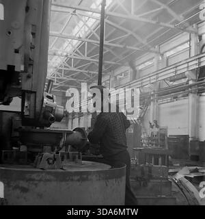 Slavyansk, Ukrainian SSR, USSR - 1970s: A young Soviet machinist in a flat cap operating a large vertical boring machine at the Stroymash factory. The worker is reaching up to adjust the spindle controls while standing on the machine platform in a sunlit workshop. This atmospheric black and white archival photo captures the concentration of youth and the scale of industrial production in the peaceful Donbass region during the Soviet era. Skilled labor on heavy equipment. Stock Photo