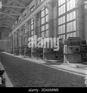 Slavyansk, Ukrainian SSR, USSR - 1970s: A wide perspective view of the machine shop interior at the Stroymash factory. A long row of heavy duty metalworking lathes is lined up along the large industrial windows, illuminating the workspace with natural light. The floor is paved with hexagonal tiles, characteristic of Soviet industrial architecture. This black and white archival photo captures the scale of manufacturing capacity and the orderly atmosphere of the peaceful Donbass region during the Soviet era. Stock Photo