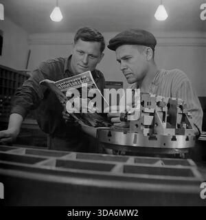 Slavyansk, Ukrainian SSR, USSR - 1970s: Two Soviet workers reading the "Inventor and Rationalizer" magazine during a break at the Stroymash factory. The men in workwear are standing next to a large metal machine part, discussing a technical article. One worker is smoking a cigarette while looking at the page. This black and white archival photo captures the thirst for knowledge and technical literacy of the working class in the peaceful Donbass region during the Soviet era. Stock Photo