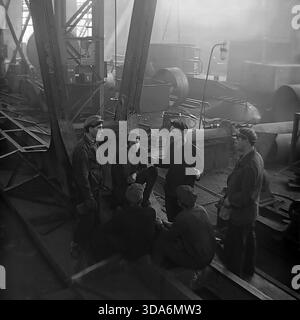 Slavyansk, Ukrainian SSR, USSR - 1970s: A group of Soviet workers having a lively conversation during a break in the assembly shop of the Stroymash factory. The men in heavy workwear and flat caps are gathered on metal structures in a smoky industrial atmosphere, discussing news or production tasks. This atmospheric black and white archival photo captures the genuine camaraderie and daily life of the working class in the peaceful Donbass region during the Soviet era. Workers meeting. Stock Photo