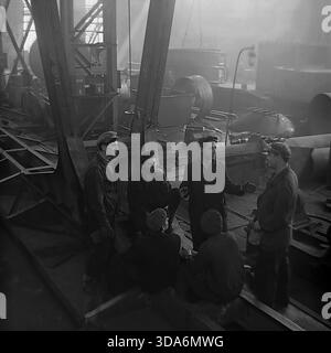 Slavyansk, Ukrainian SSR, USSR - 1970s: A group of Soviet workers having a lively conversation during a break in the assembly shop of the Stroymash factory. The men in heavy workwear and flat caps are gathered on metal structures in a smoky industrial atmosphere, discussing news or production tasks. This atmospheric black and white archival photo captures the genuine camaraderie and daily life of the working class in the peaceful Donbass region during the Soviet era. Workers meeting. Stock Photo