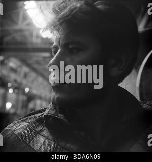 Slavyansk, Ukrainian SSR, USSR - 1970s: A cinematic close-up profile portrait of a young Soviet factory worker in the Stroymash plant workshop. The handsome man in a plaid shirt is looking into the distance, illuminated by dramatic natural light coming from the roof skylights. The background features blurred industrial structures. This atmospheric black and white archival photo captures the human face, determination and spirit of the working class in the peaceful Donbass region during the Soviet era. Stock Photo
