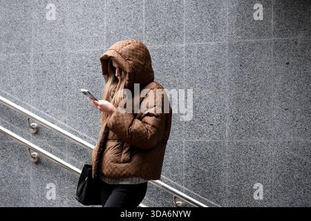 A young woman dressed in a cozy brown coat looks at her phone, walking down stairs. Kyiv, Ukraine. December 08, 2025. Stock Photo