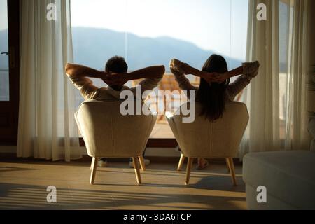 Back view of a couple relaxing at home looking through a window sitting on armchairs Stock Photo