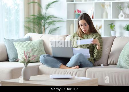Perplexed woman in green reading letter with laptop sitting on a sofa at home Stock Photo