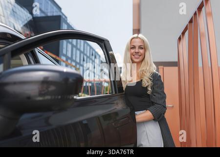 A woman with long hair is next to a black car parked by modern buildings. Stock Photo
