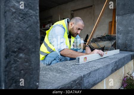 A construction worker wearing a reflective safety vest uses a spirit level to check the alignment of a stone surface. Stock Photo