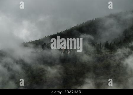 View of a building shrouded in mist, amidst a dense forest clinging to the steep slopes under a brooding sky, evoking mystery and serene isolation, Pa Stock Photo